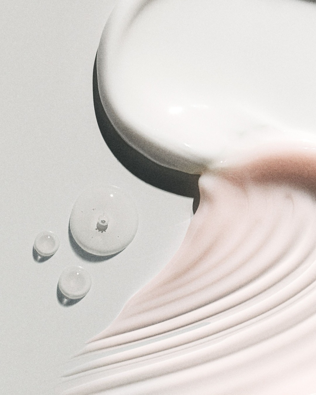 Nahaufnahme von Hautpflegeprodukten: cremeweiße und rosafarbene Texturen sowie klare Tropfen auf hellem Untergrund.