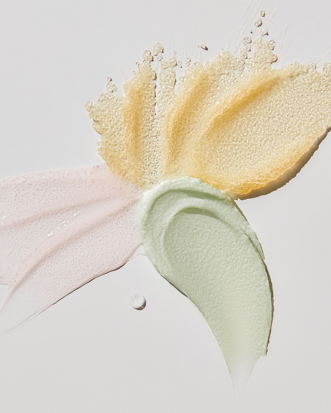 Na białym tle widoczne są próbki kosmetyków: złoty peeling z drobinkami, różowy krem o lekkiej konsystencji i zielona maska o gładkiej teksturze.