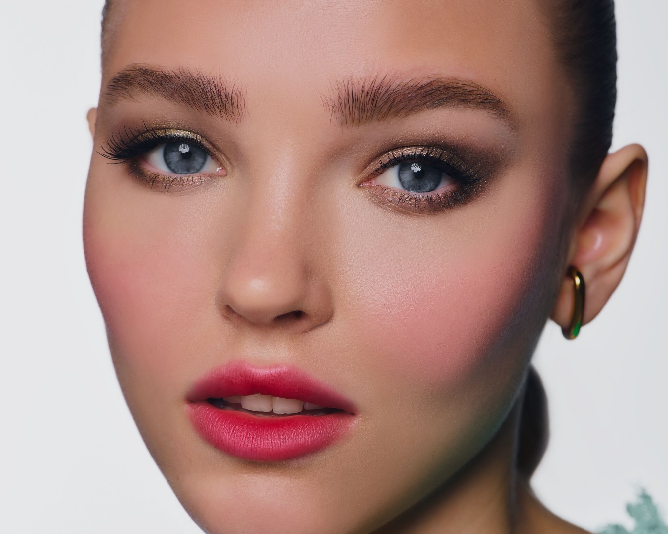 Nahaufnahme eines Frauengesichts mit strahlend blau-grauen Augen, betont durch goldenen Lidschatten und langen Wimpern, dazu rosige Wangen und leuchtend rote Lippen.
