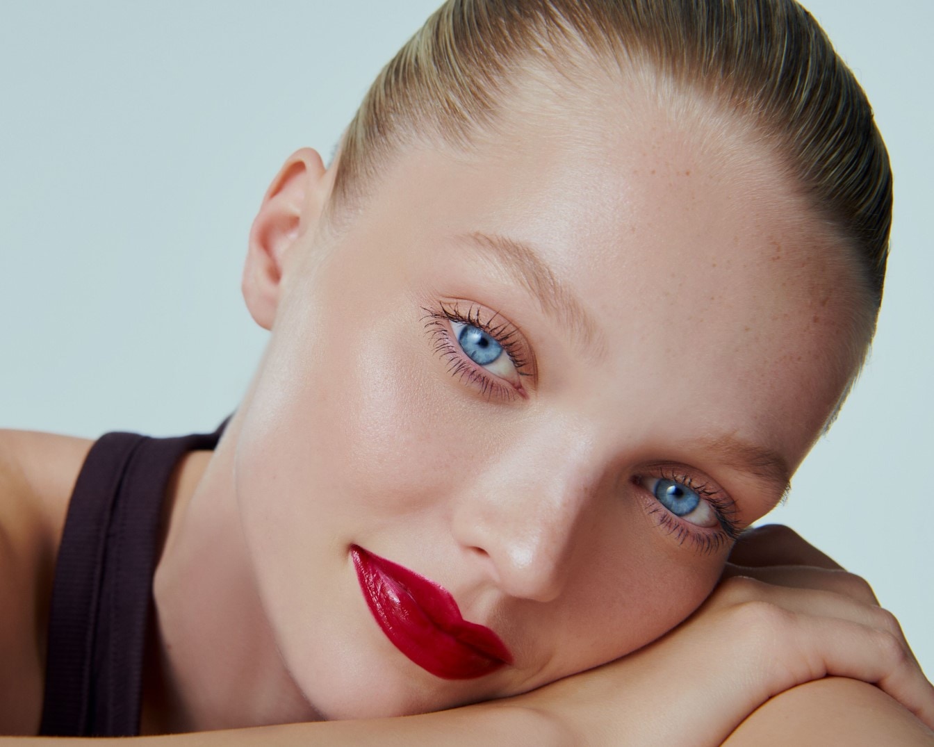 Een close-up van een vrouw met blauwe ogen en rode lippenstift, haar blonde haar strak naar achteren gekamd, leunend op haar armen.