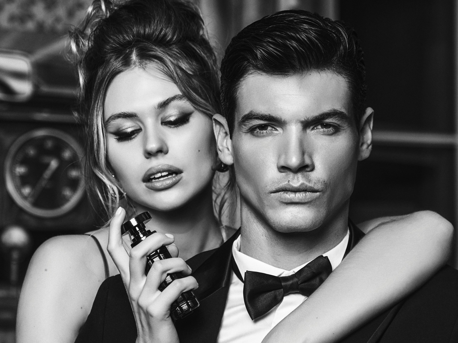 Primo piano in bianco e nero di una donna che spruzza profumo su un uomo in smoking con papillon, creando un'atmosfera elegante e sofisticata.