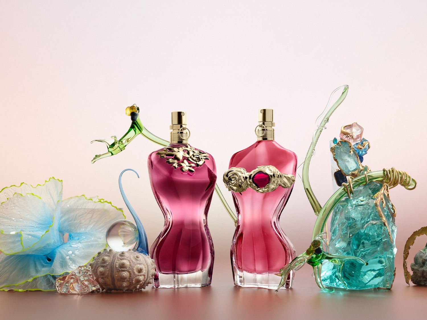 Twee parfumflessen in de vorm van een vrouwelijk torso, de ene in donkerroze met gouden bloemmotieven en de andere in lichtroze met een gouden roos, staan tussen decoratieve glazen en stenen elementen.