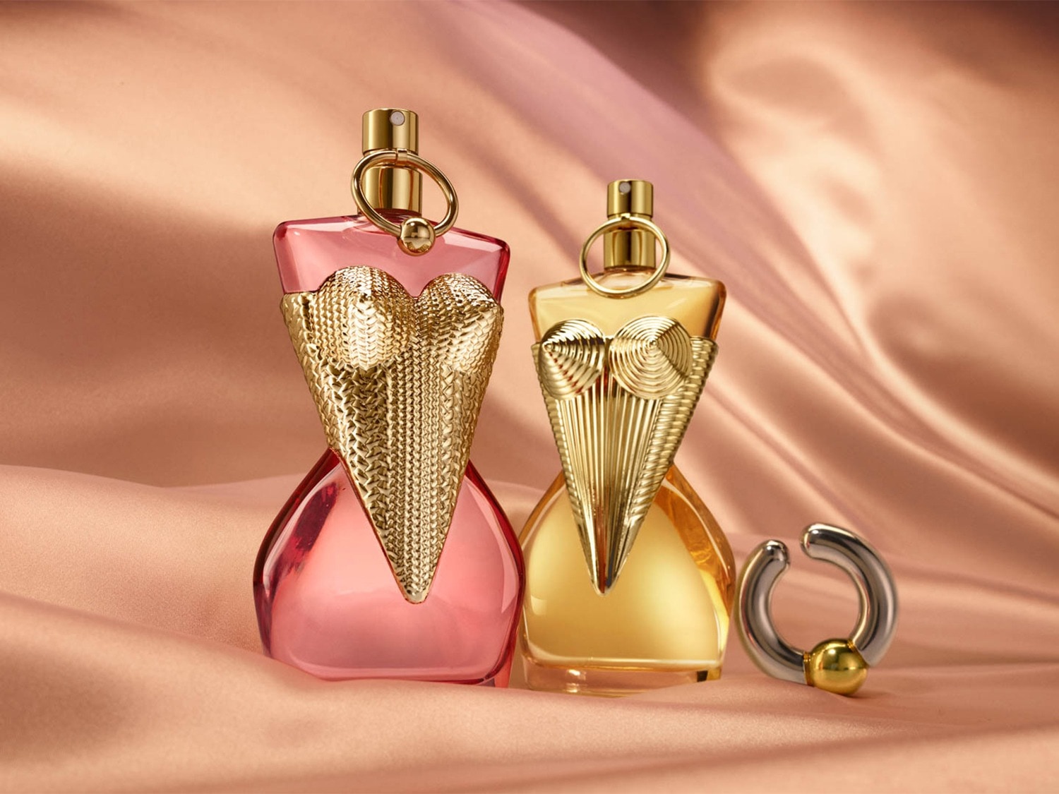 Twee parfumflesjes in de vorm van een vrouwelijk torso, één roze en één goudkleurig, staan op een glanzende perzikkleurige stof, met een zilveren en gouden oorbel ernaast.