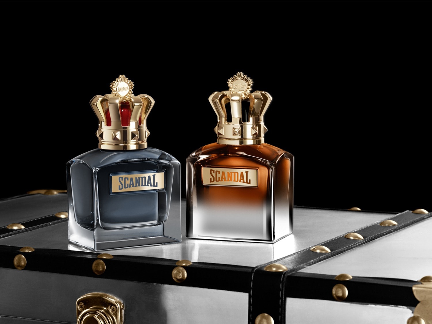 Twee flacons van Scandal parfum, één met een donkerblauwe vloeistof en de andere met een amberkleurige vloeistof, beide met een gouden kroonvormige dop, staan op een glanzende koffer met gouden accenten.