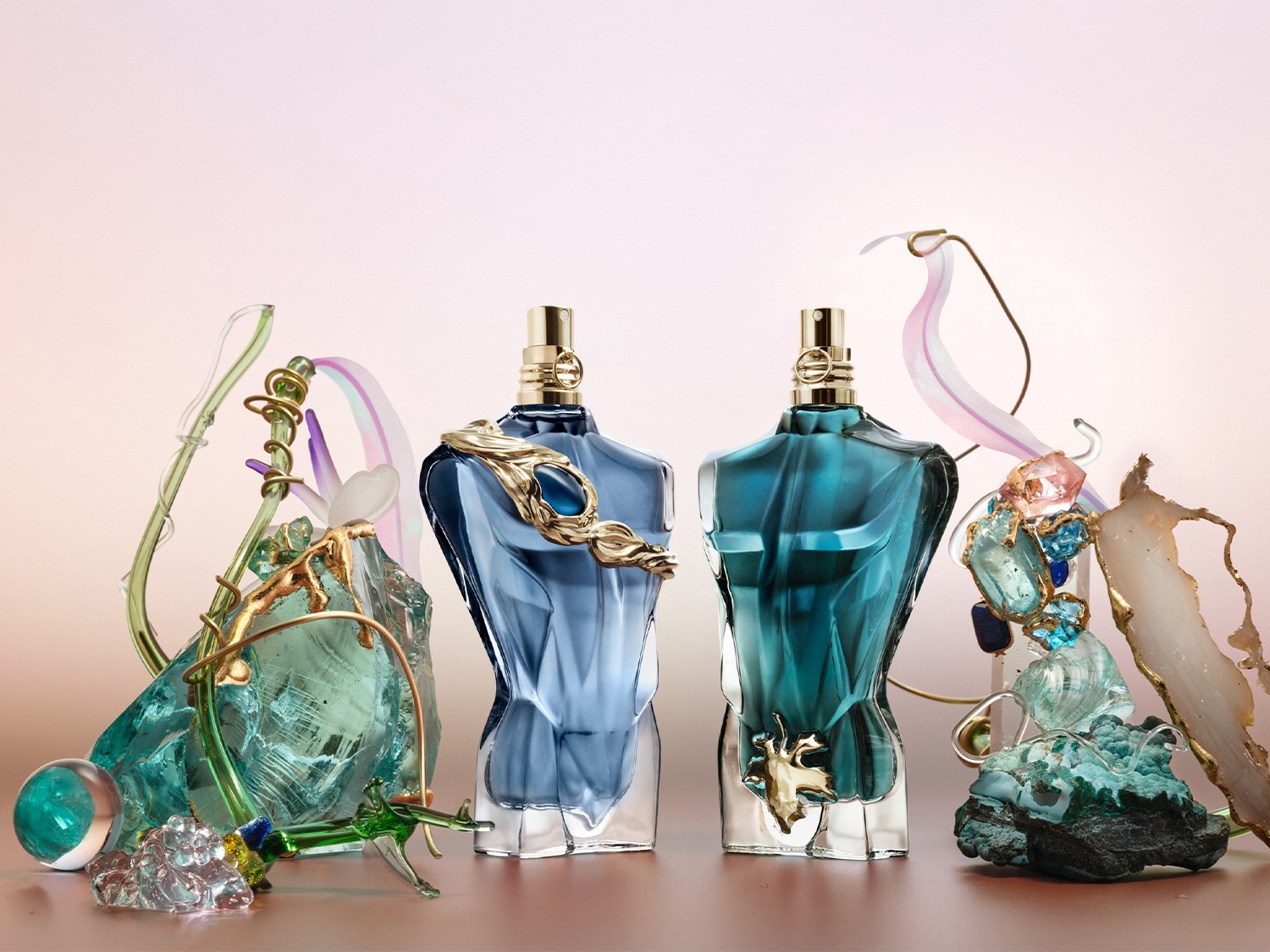 Twee Jean Paul Gaultier Le Male parfums in de vorm van een mannentorso, de linker in lichtblauw en de rechter in donkerblauw, staan tussen abstracte, kleurrijke glazen sculpturen.