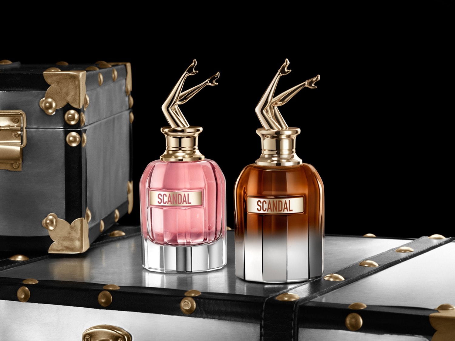 Twee flacons van Jean Paul Gaultier Scandal parfum staan op een koffer, de linker is roze en de rechter is bruin, beide met een gouden dop in de vorm van benen.