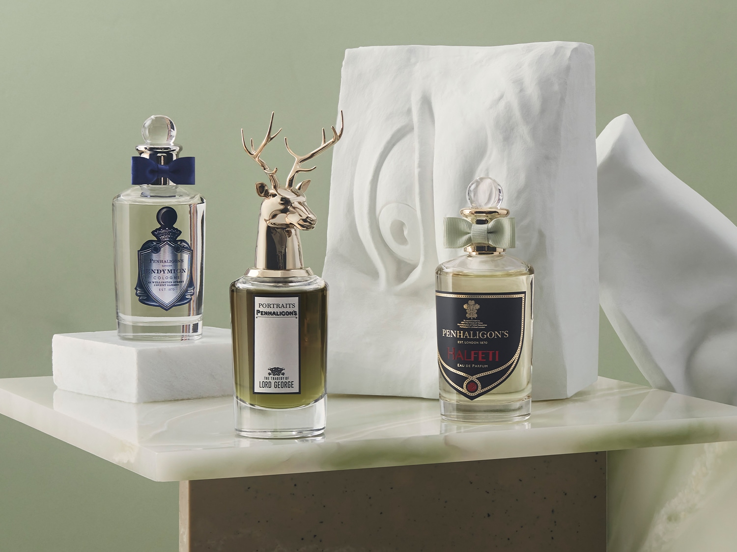 Drei Penhaligon's Parfümflakons stehen auf einem Marmortisch, darunter Endymion Cologne mit blauer Schleife, Lord George mit goldenem Hirschkopf und Halfeti Eau de Parfum mit grüner Schleife.