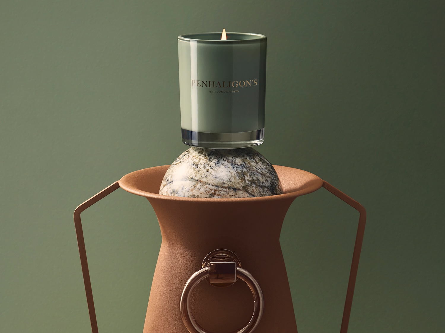 Eine grüne Penhaligon's Duftkerze steht auf einem Marmorstein, der in einer braunen Vase platziert ist und eine warme, einladende Atmosphäre schafft.