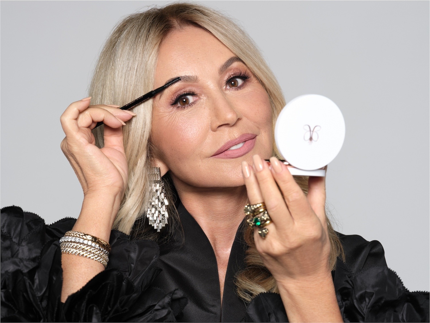 Blondwłosa kobieta w czarnej bluzce, z kolczykami i pierścionkami, używa pędzelka do brwi, trzymając w drugiej ręce lusterko.