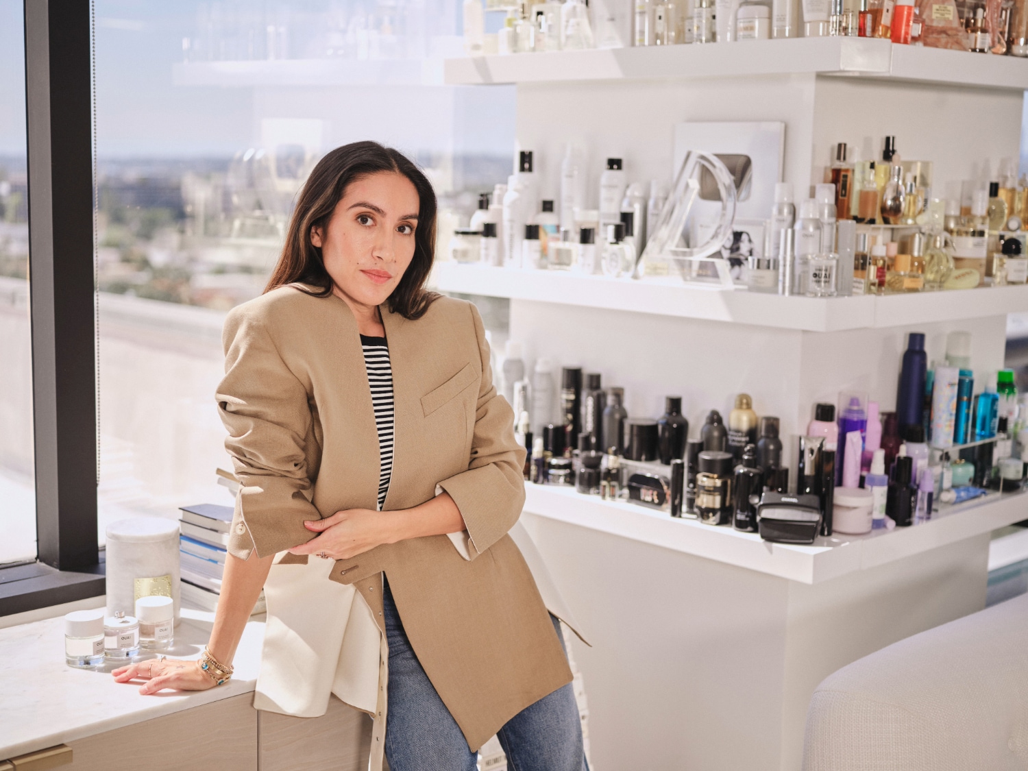 Kobieta w beżowej marynarce opiera się o marmurowy blat, na którym stoją białe słoiczki z kosmetykami, a w tle widać półki uginające się od produktów do pielęgnacji.