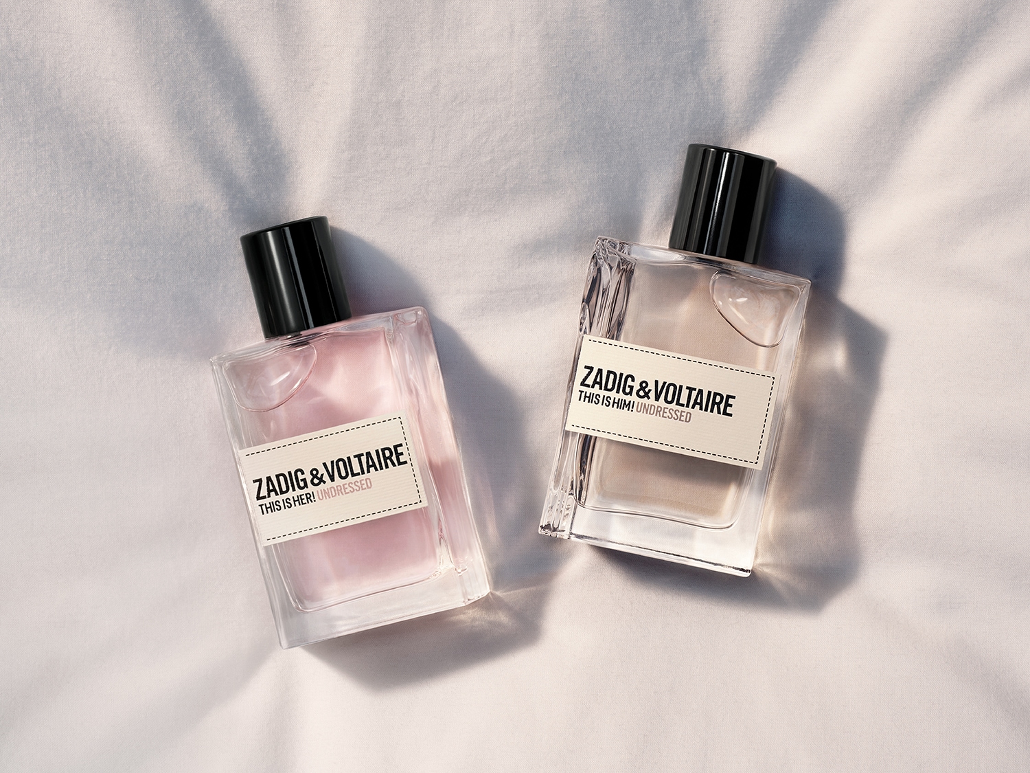 Zwei Parfümflaschen von Zadig & Voltaire liegen auf einem weißen Tuch, einmal "This is her! Undressed" mit rosa Flüssigkeit und einmal "This is him! Undressed" mit beiger Flüssigkeit.