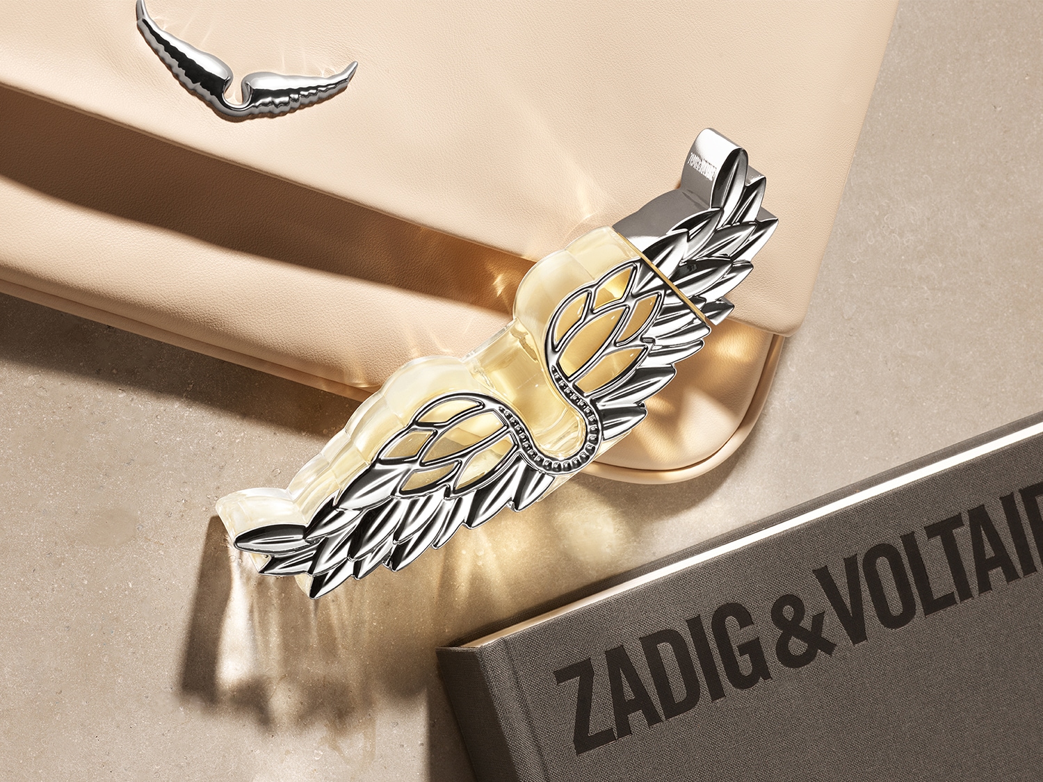 Nahaufnahme des Zadig & Voltaire Parfüms mit silbernen Flügeln, das auf einer beigen Handtasche und einem Buch mit dem Aufdruck 'ZADIG & VOLTAIRE' liegt.