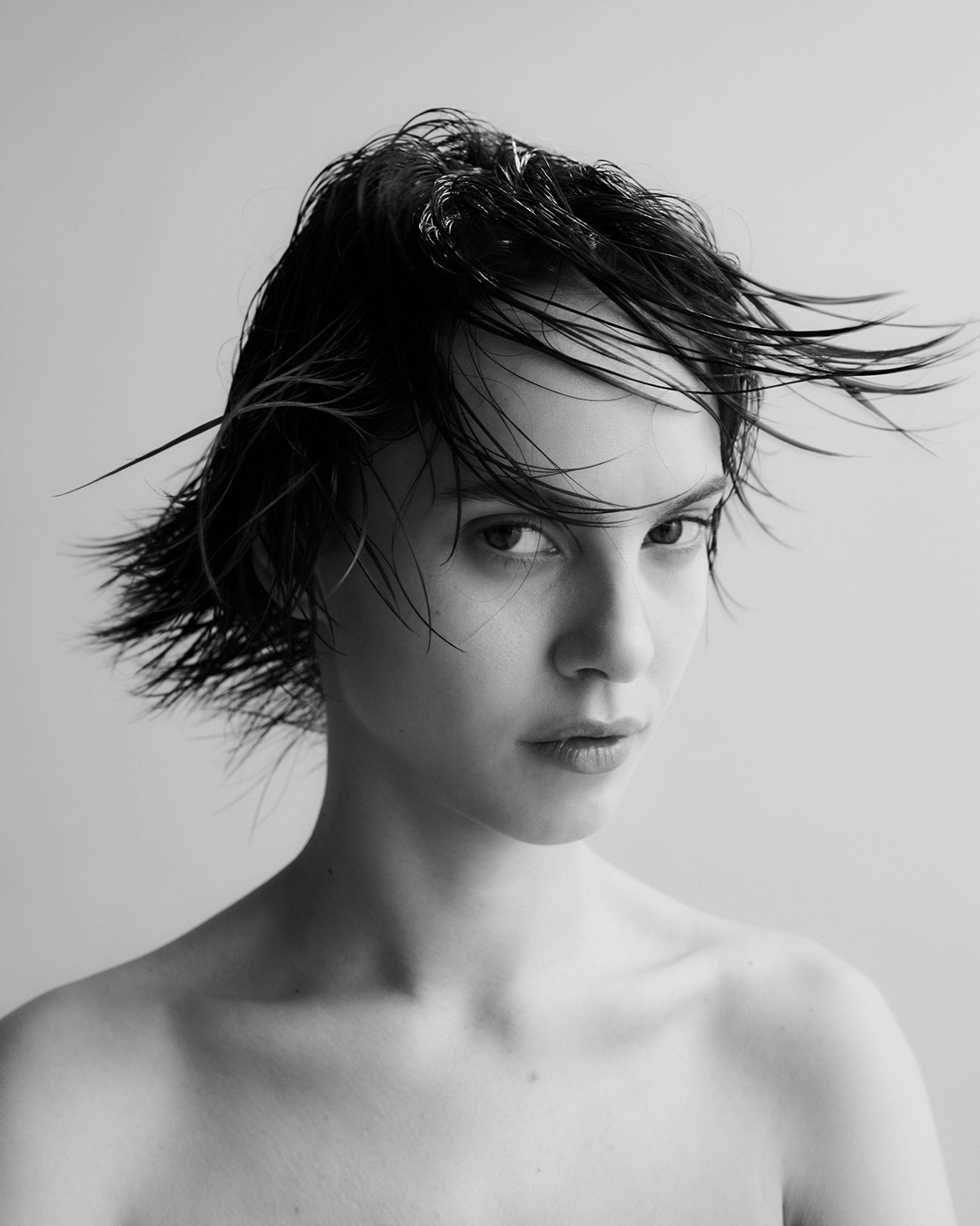 Czarno-białe zdjęcie kobiety z krótkimi, mokrymi włosami, skierowanej wzrokiem prosto w obiektyw, na jasnym tle.