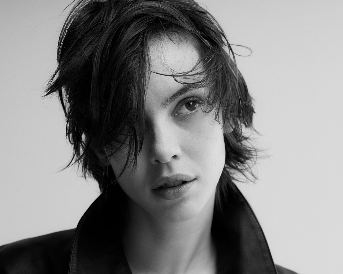 Schwarz-Weiß-Porträt einer Frau mit dunklen, zerzausten Haaren und einem entschlossenen Blick, die eine Lederjacke mit breitem Kragen trägt.