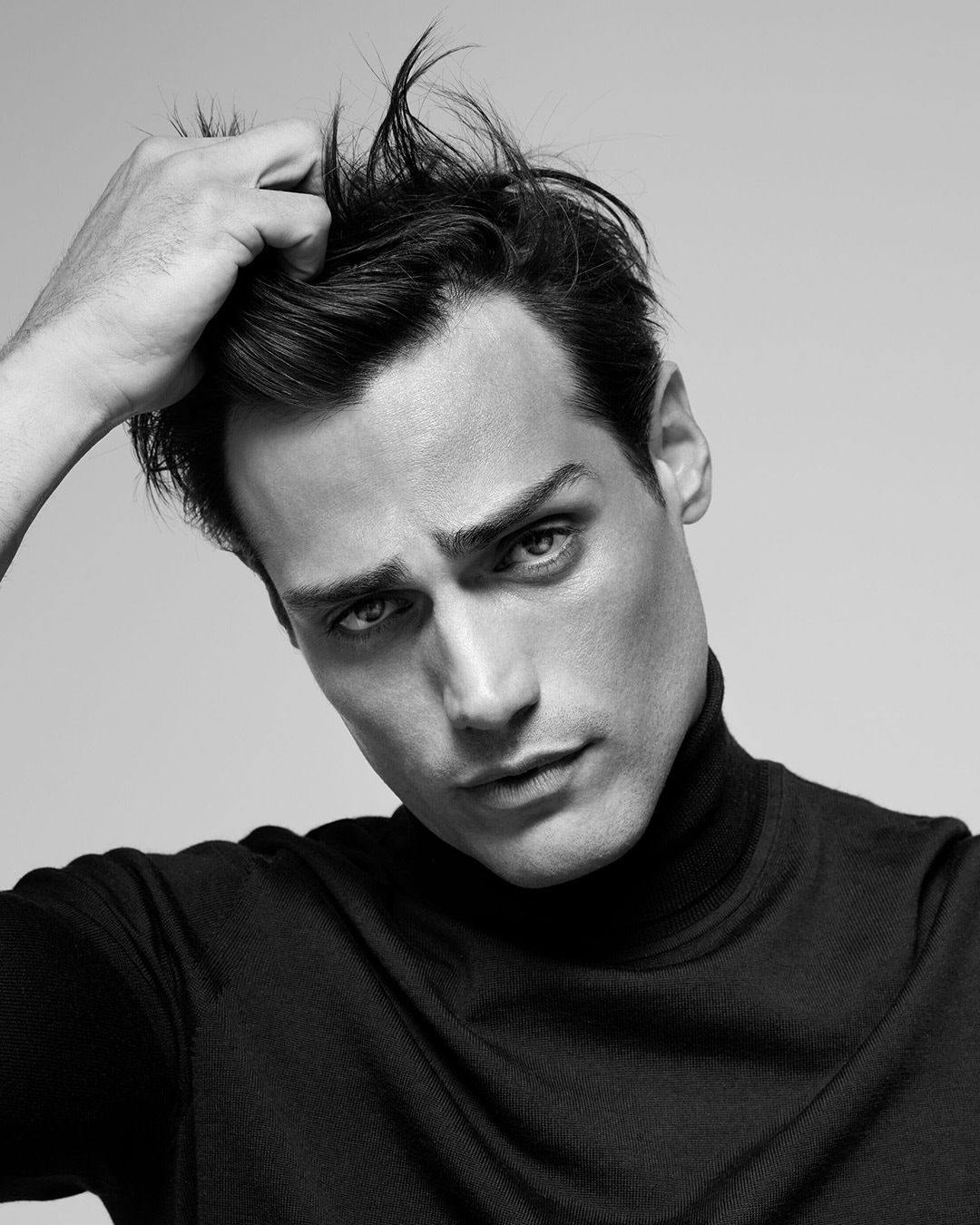 Czarno-białe ujęcie mężczyzny w czarnym golfie, który nonszalancko poprawia włosy, spoglądając wprost w obiektyw.