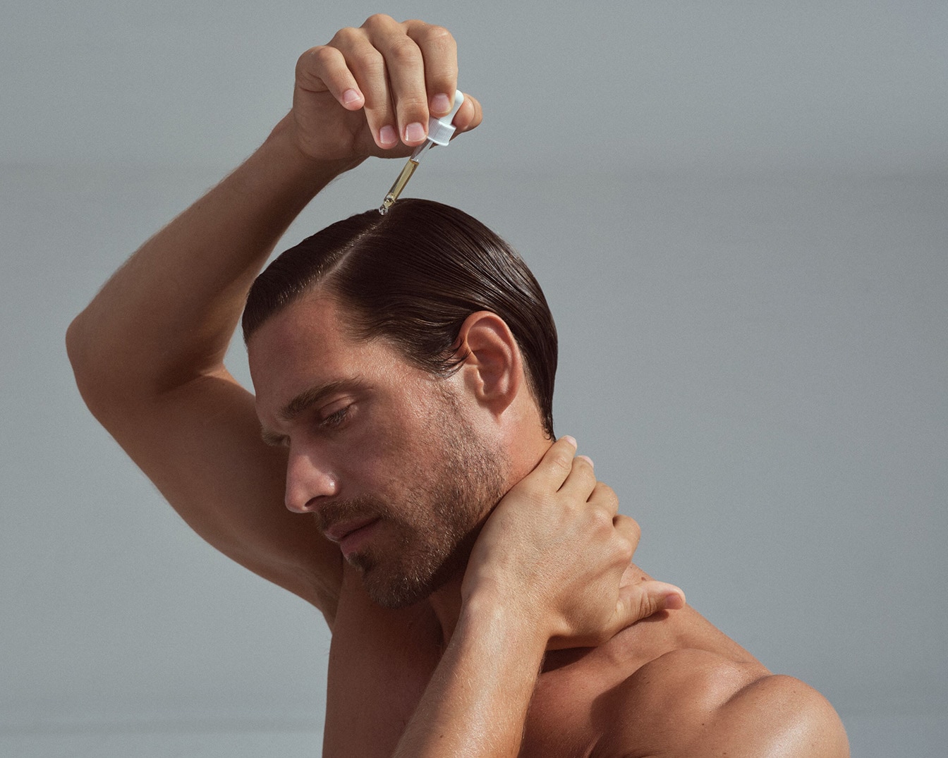 Ein Mann mit nacktem Oberkörper massiert sich Öl mit einer Pipette in sein glänzendes, zurückgekämmtes, braunes Haar.