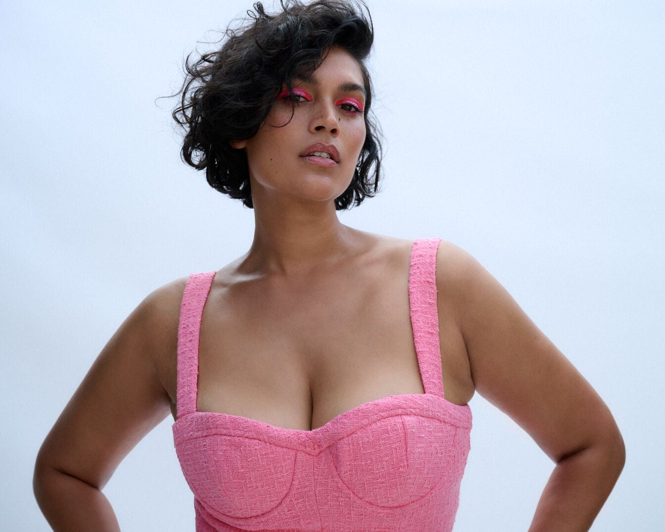 Moters portretas su rožiniais akių šešėliais ir rožine suknele, pozuojanti rankomis ant klubų.