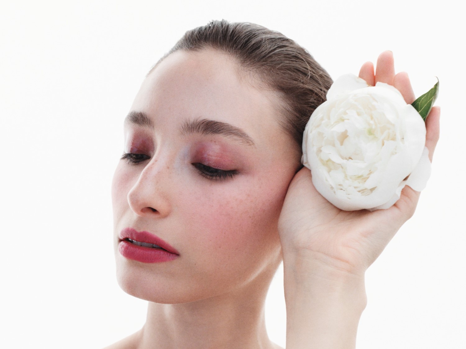Eine Frau mit geschlossenen Augen hält eine weiße Blume an ihr Gesicht, ihr Make-up betont ihre rosigen Wangen und Lippen.