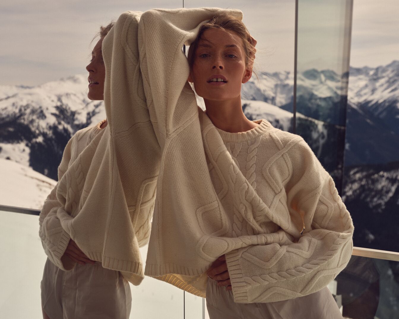 Eine Frau in einem cremefarbenen Strickpullover posiert vor einer Bergkulisse, ihr Spiegelbild ist sichtbar.