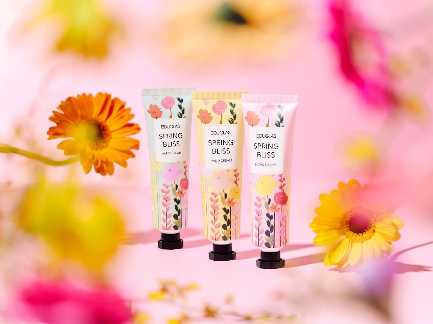 Tri krémy na ruky Douglas Spring Bliss s kvetinovým dizajnom v ružovej, žltej a zelenej farbe, obklopené kvetmi, vytvárajúce jarnú atmosféru.