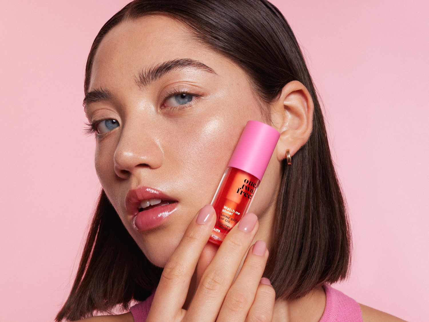 Na różowym tle, kobieta prezentuje błyszczący olejek do ust One Two Free w przezroczystej buteleczce z różową zakrętką, który obiecuje efekt "shine bright".