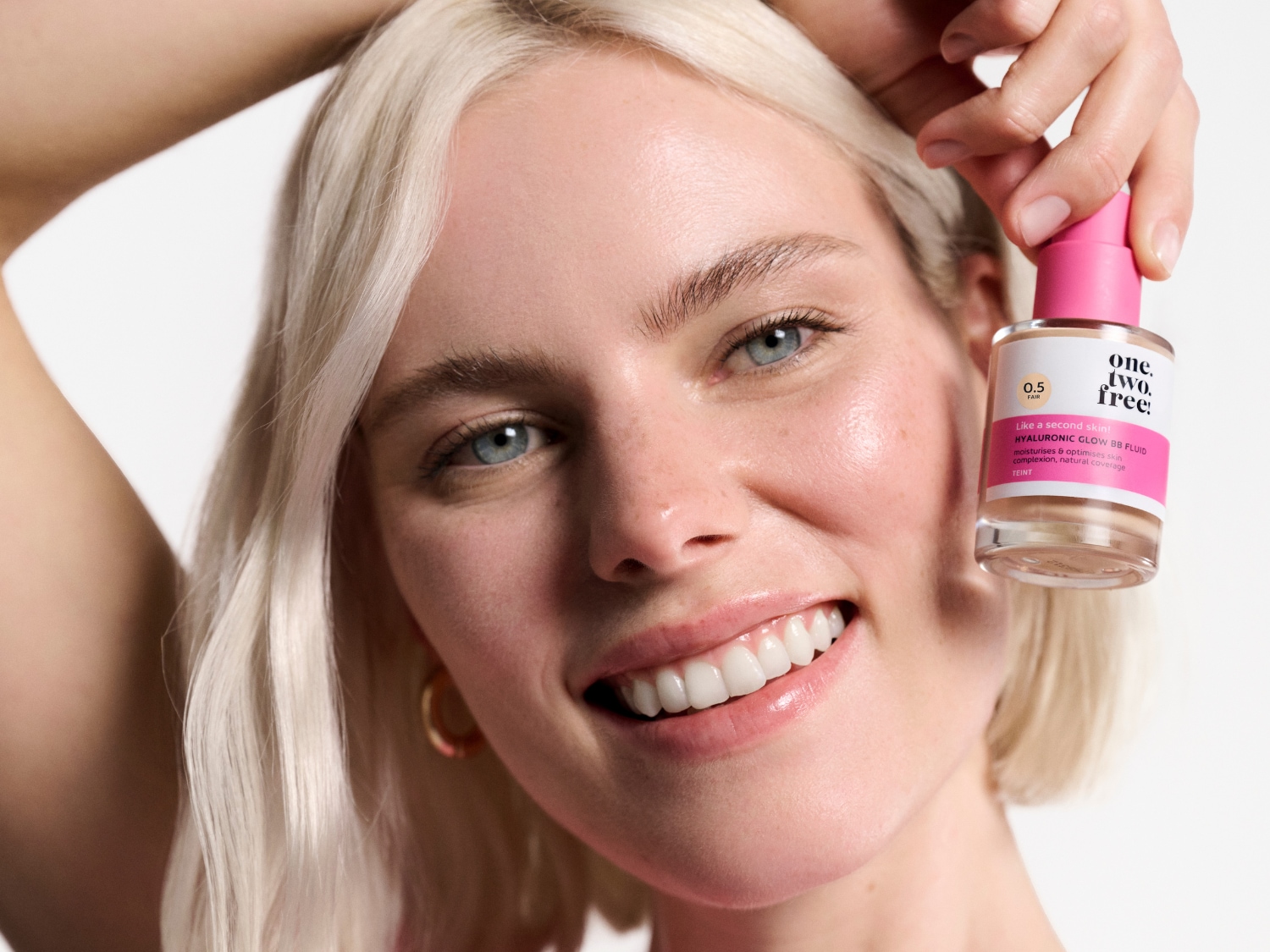 Uśmiechnięta blondwłosa kobieta trzymająca w dłoni przezroczystą butelkę BB fluidu Hyaluronic Glow z różową zakrętką, który nawilża, poprawia koloryt i zapewnia naturalne krycie.