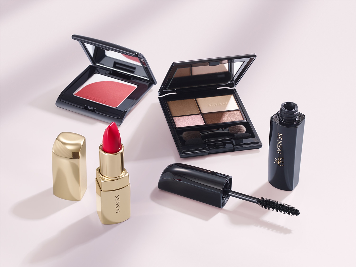 Ontdek de Sensai make-up collectie: een rouge met spiegel in een zwart doosje, een oogschaduw palette met kwast, een rode lipstick in een gouden huls en een zwarte mascara.