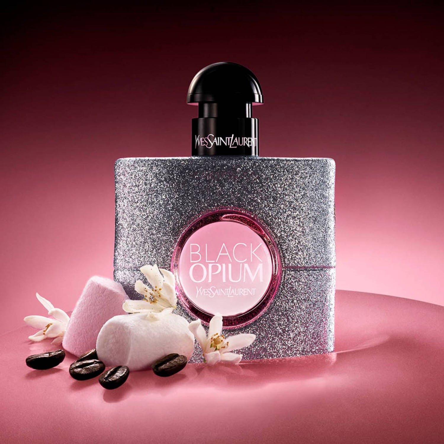A Black Opium Yves Saint Laurent parfümös üveg csillogó ezüstben, fekete kupakkal, rózsaszín logóval, kávészemekkel, fehér virágokkal és rózsaszín cukorkákkal.