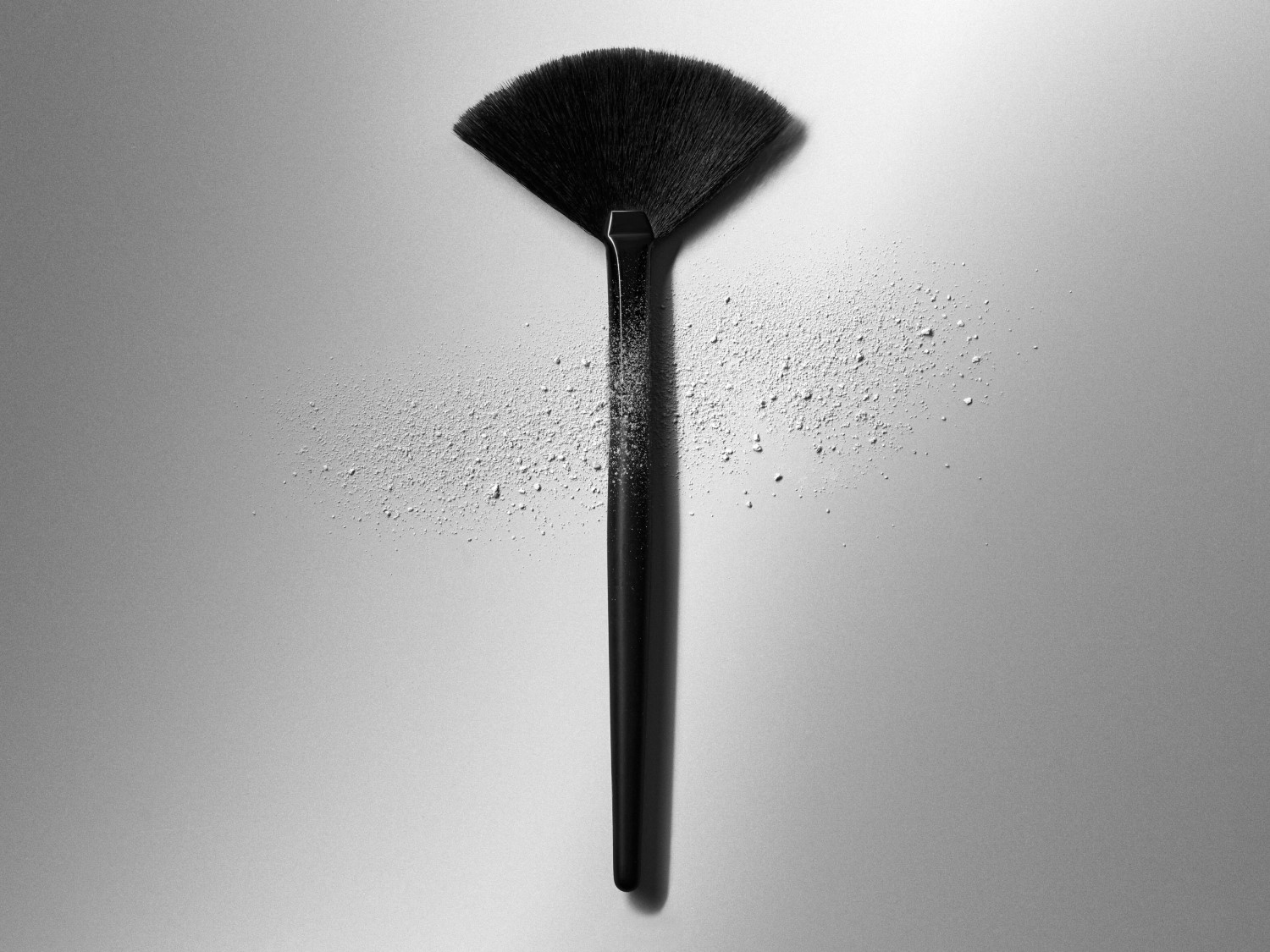 Ein schwarzer Fächerpinsel liegt auf einem grauen Untergrund, umgeben von Puderpartikeln, ideal zum Auftragen von Make-up für ein ebenmäßiges Finish.