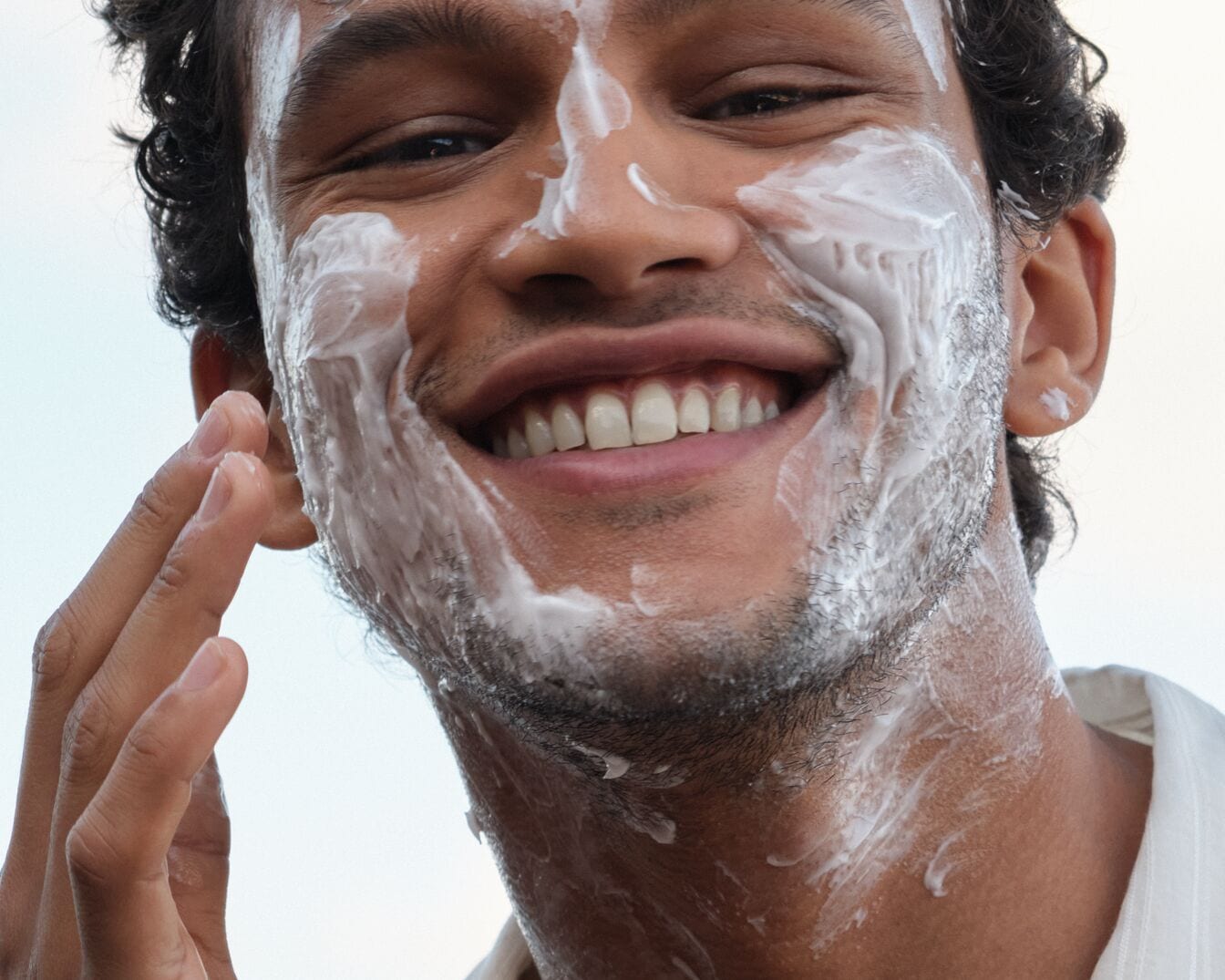 Uśmiechnięty mężczyzna z kręconymi włosami nakłada biały krem na twarz, delikatnie wklepując go opuszkami palców.
