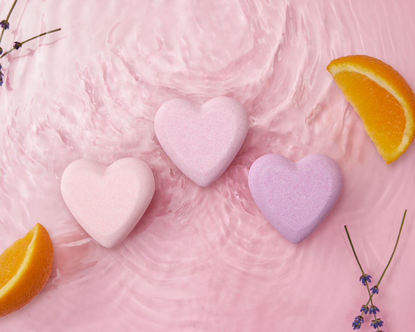 Trzy serca do kąpieli w odcieniach różu i fioletu unoszą się na różowej wodzie, otoczone plasterkami pomarańczy i gałązkami lawendy, tworząc relaksującą i aromatyczną scenerię.