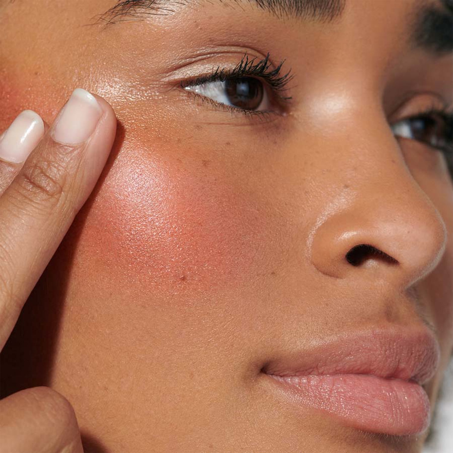 Nahaufnahme eines Gesichtes mit einem Hauch von Rouge auf den Wangen, der mit einem Finger aufgetragen wird, um einen strahlenden Teint zu erzielen.