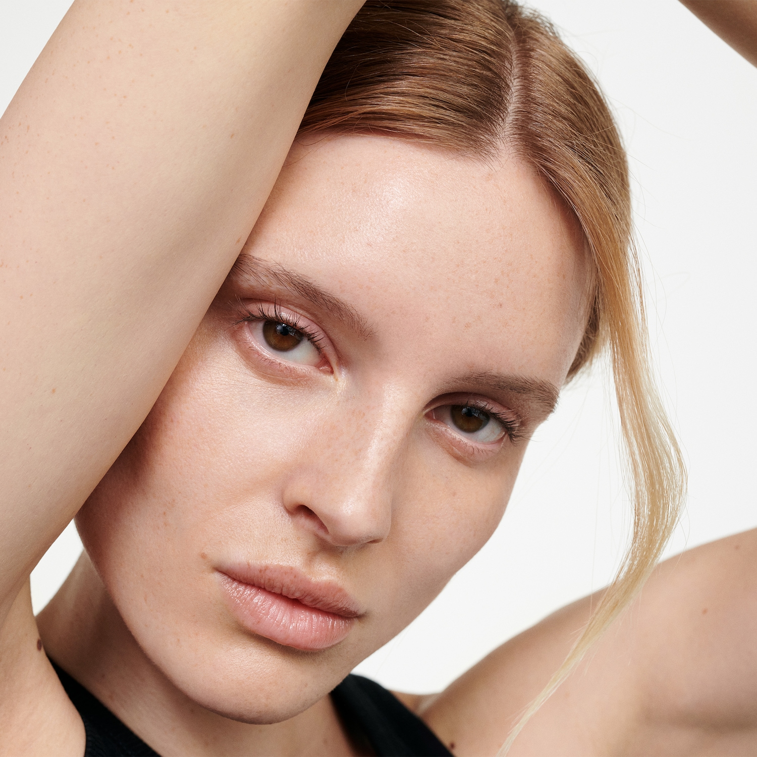 Nahaufnahme einer Frau mit hellbraunen Augen und blonden Haaren, die ihre Arme über den Kopf hält, wodurch ihre natürliche Schönheit und ihr Selbstbewusstsein betont werden.