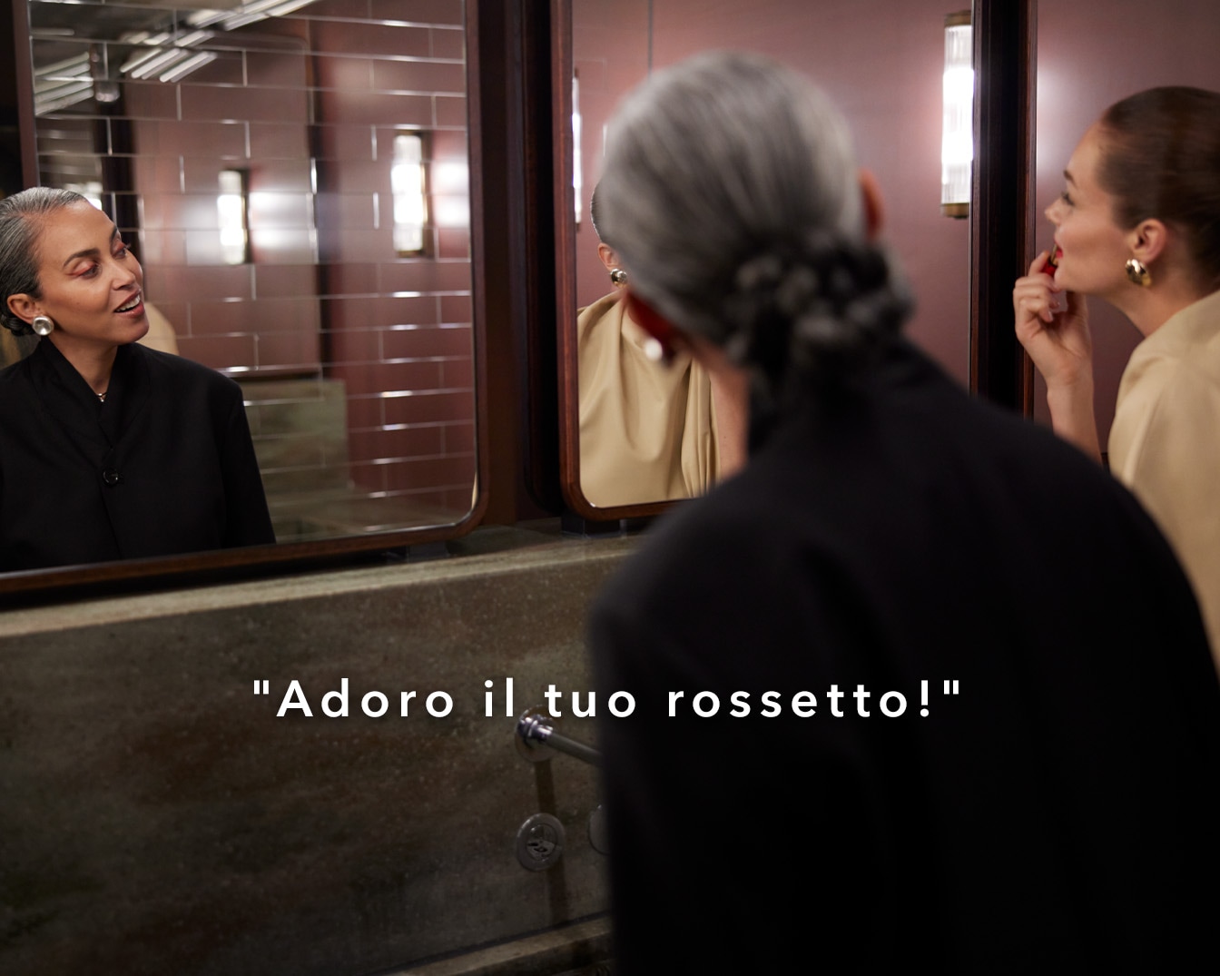 Two women in a bathroom, one admires the other's red lipstick in the mirror, saying "Adoro il tuo rossetto!"