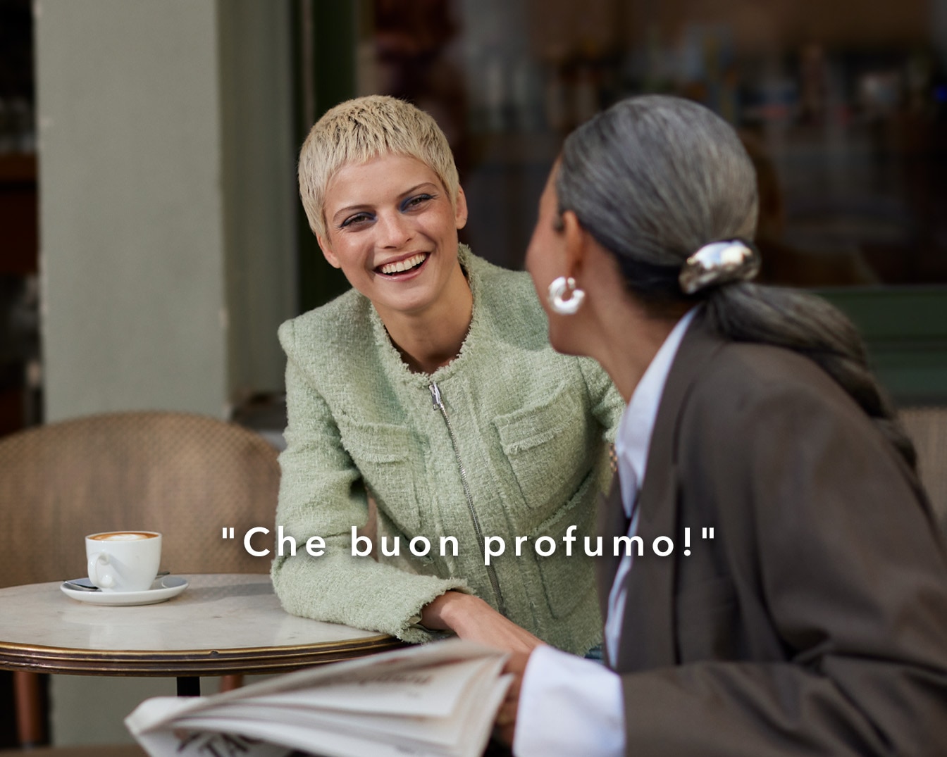 Due donne sedute a un tavolo di caffè, una bionda con una giacca verde sorride mentre l'altra, con i capelli grigi raccolti, indossa un blazer marrone; si percepisce un'atmosfera amichevole e rilassata.