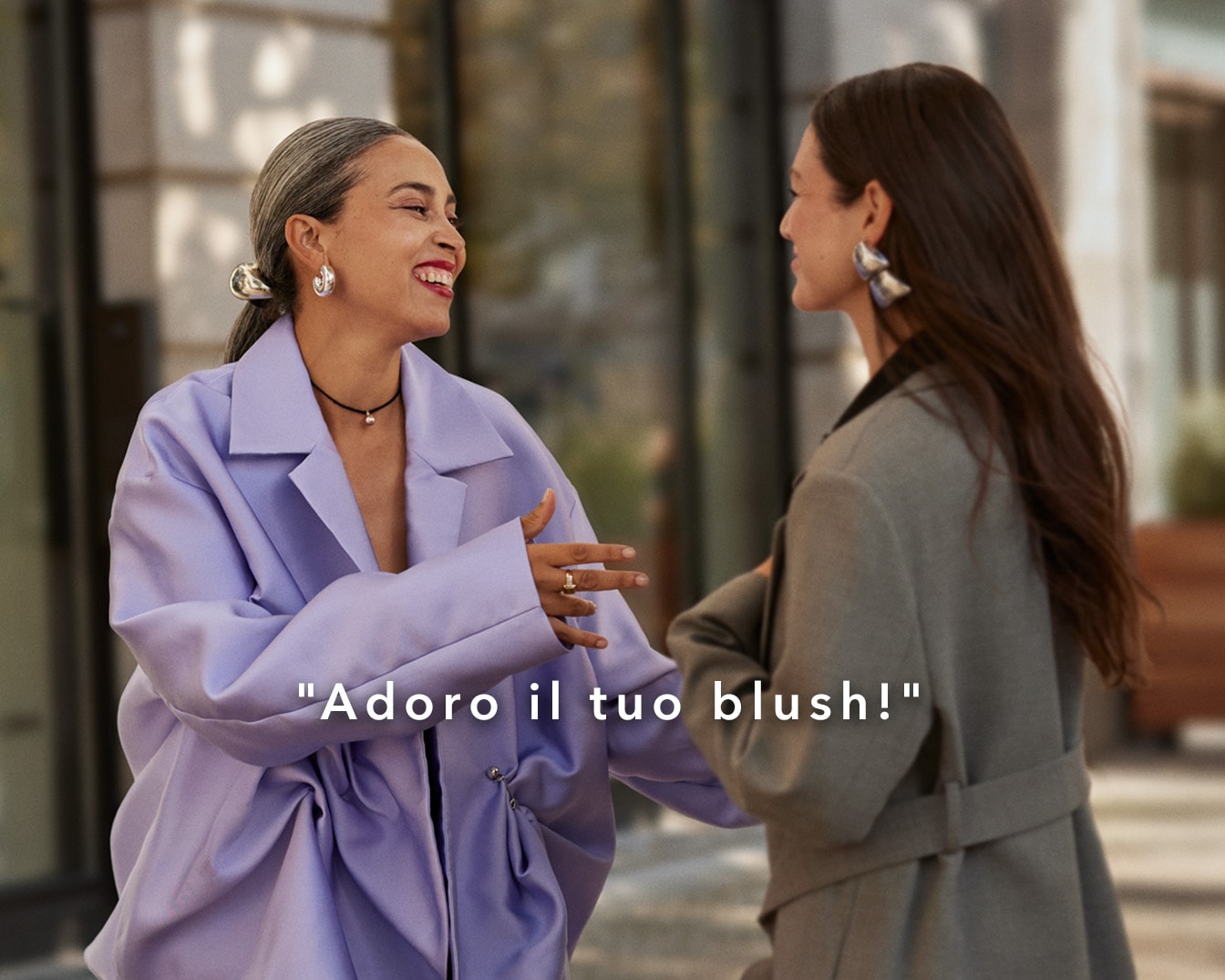 Due donne sorridono mentre una, con un cappotto lilla, esclama "Adoro il tuo blush!", mettendo in risalto un momento di apprezzamento per il trucco.
