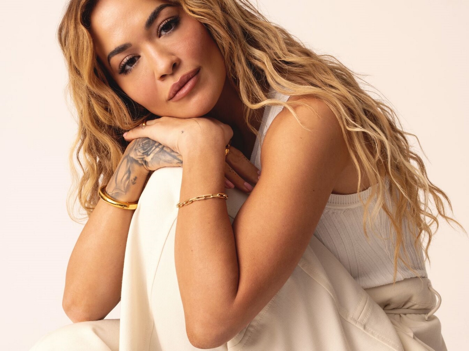 Portrait de Rita Ora, les cheveux ondulés et blonds, portant un débardeur blanc et des bijoux dorés, les mains croisées sous le menton.