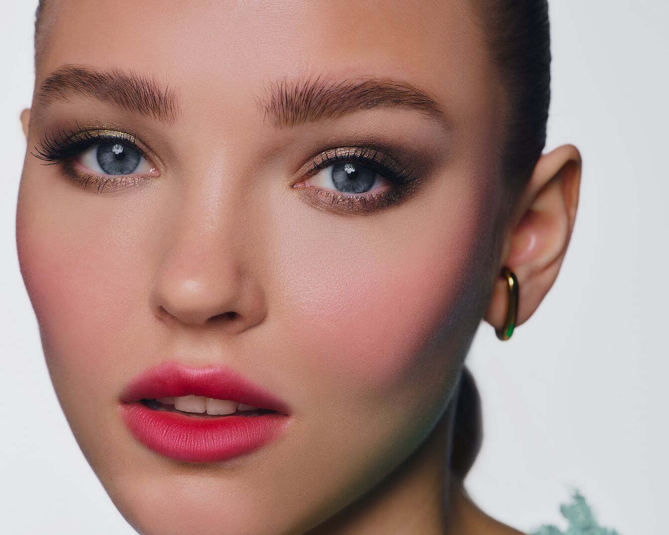 Nahaufnahme eines Frauengesichts mit strahlend blau-grauen Augen, betont durch schimmernden Gold- und Brauntönen im Lidschatten, rosigen Wangen und einem Hauch von rotem Lippenstift.