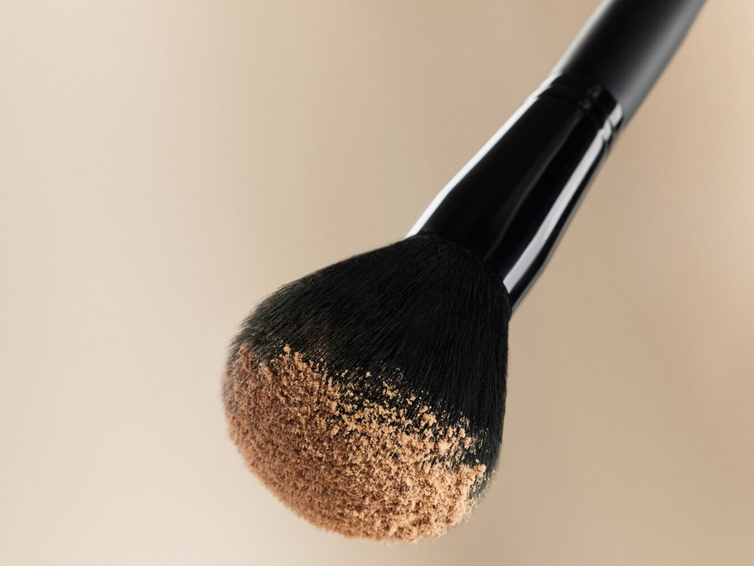 Aquí se muestra una brocha de maquillaje con mango negro y cerdas suaves, impregnada de polvos sueltos de color beige.