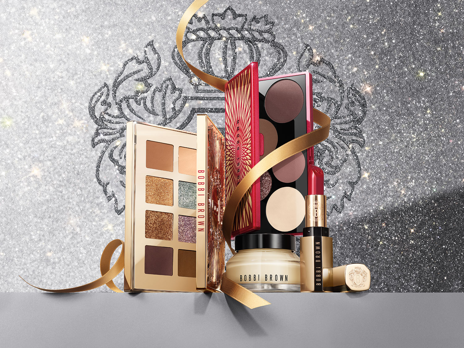 A Bobbi Brown sminktermékekből álló kollekció arany szalaggal átkötve, csillogó ezüst háttér előtt, melyben szemhéjpúder paletták, rúzs és arckrém is található.