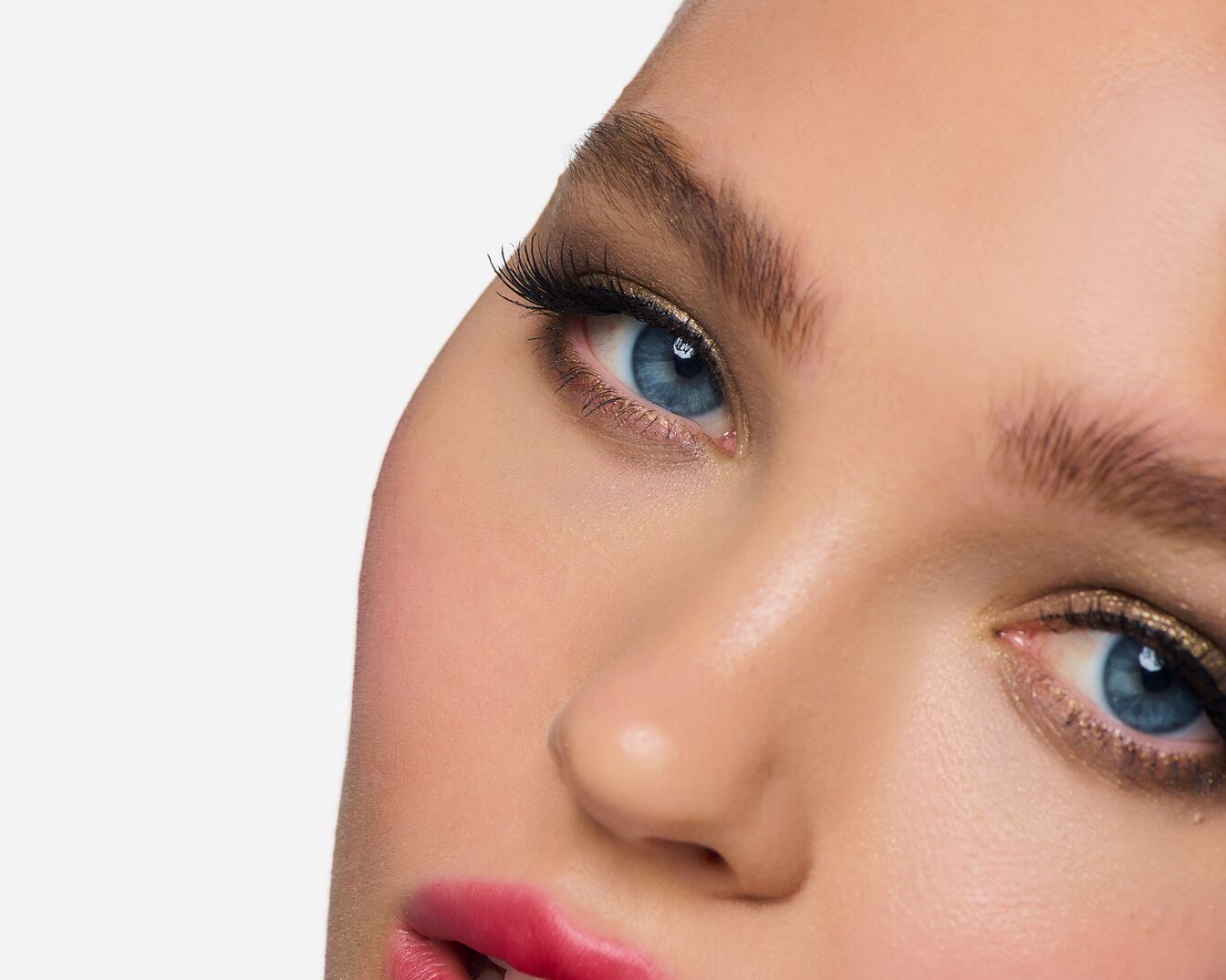 Close-up van een gezicht met blauwe ogen, gouden oogschaduw, lange wimpers, roze lippen en een heldere huid.