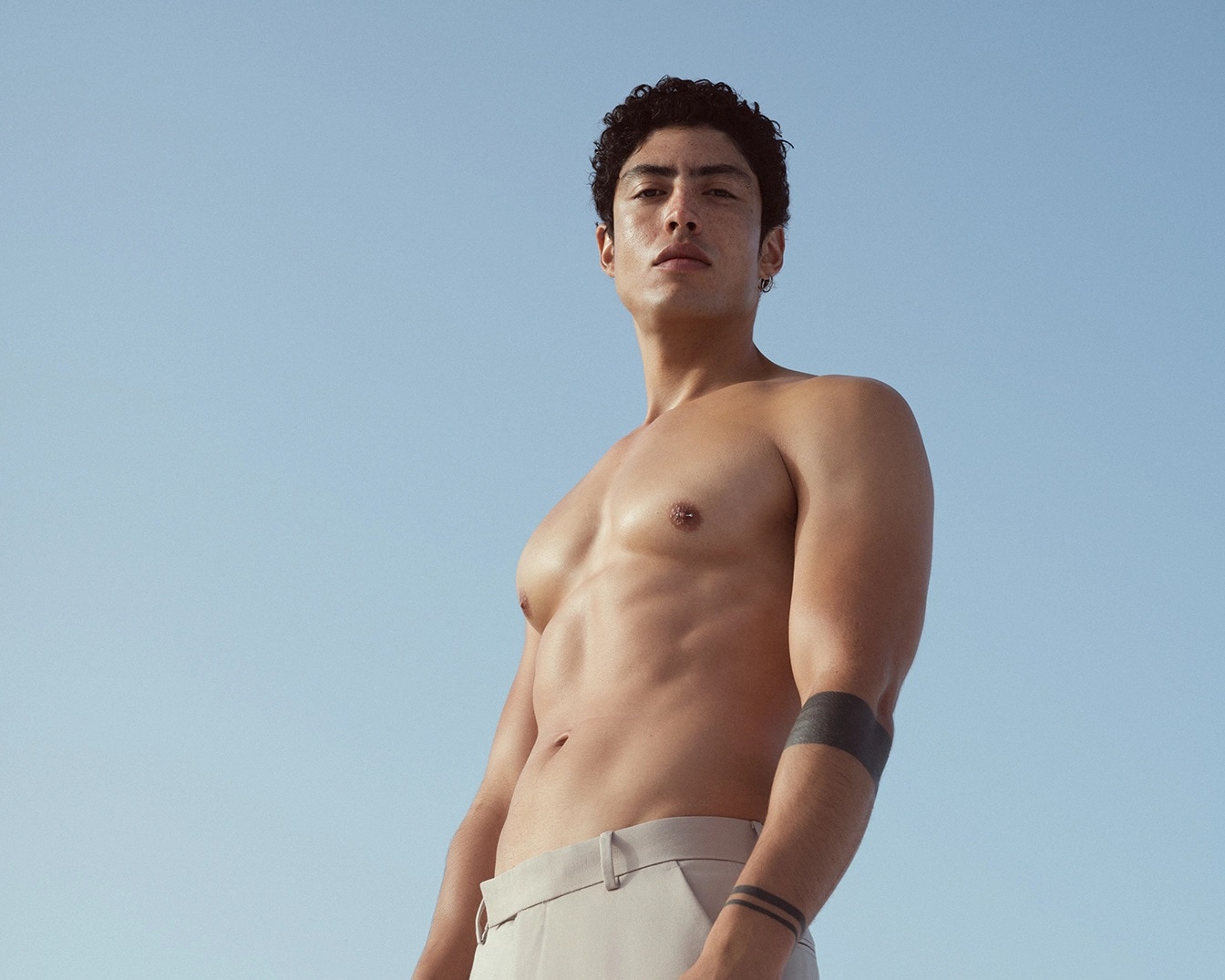 Uomo muscoloso con i capelli ricci, la pelle abbronzata e pantaloni color crema, che mostra un tatuaggio nero sul braccio contro un cielo azzurro.