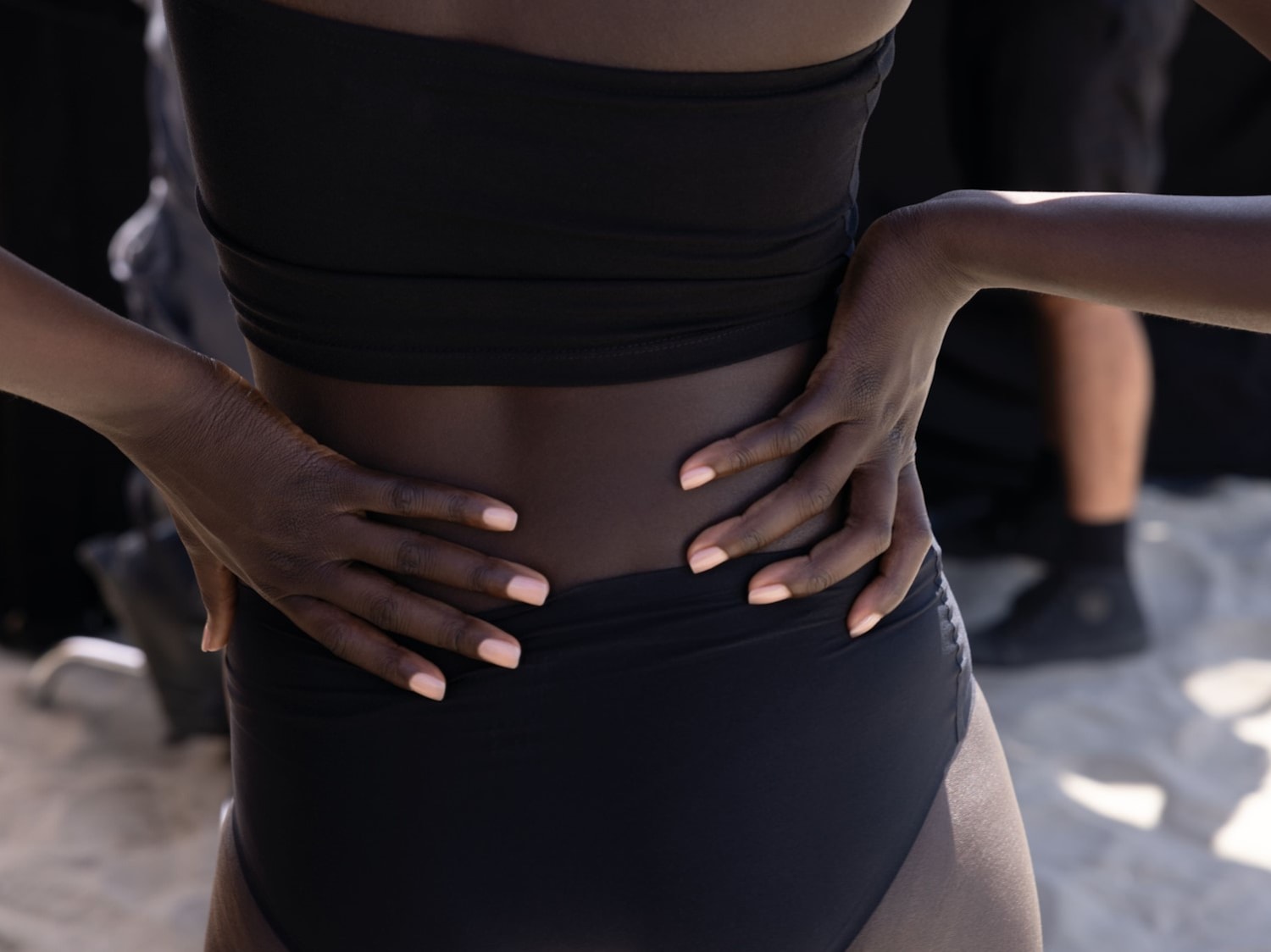 Een vrouw met een donkere huidskleur draagt een zwarte top en broek, haar handen rusten op haar heupen.