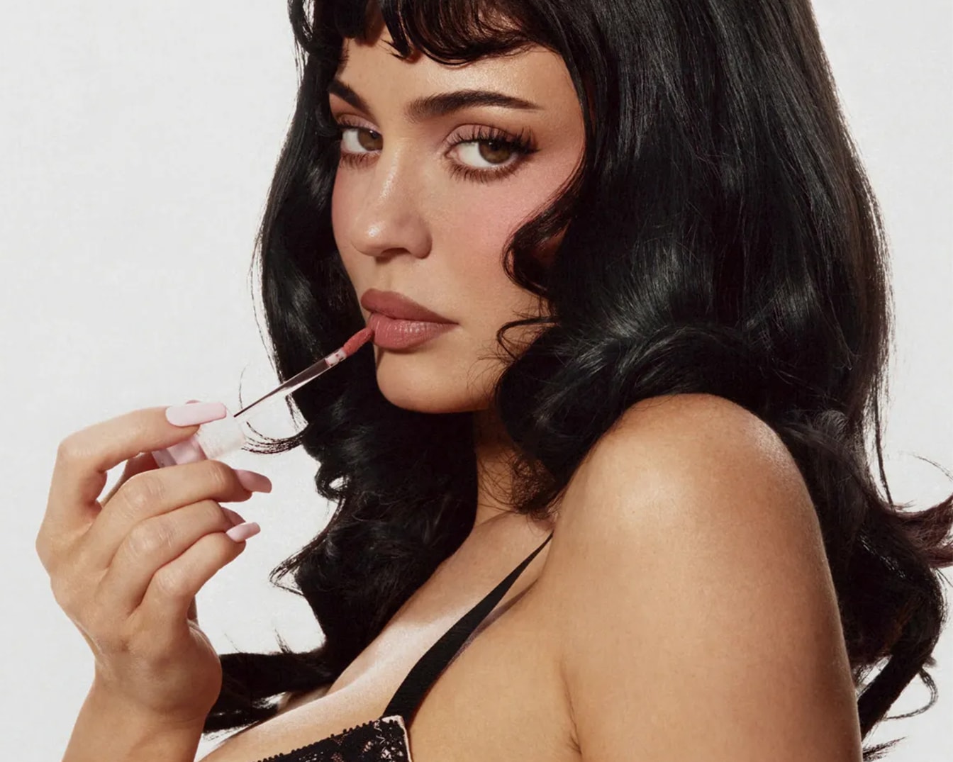 Una donna con capelli scuri e mossi applica un lucidalabbra nude con un applicatore, mettendo in risalto le sue labbra carnose e le unghie lunghe e rosa chiaro.