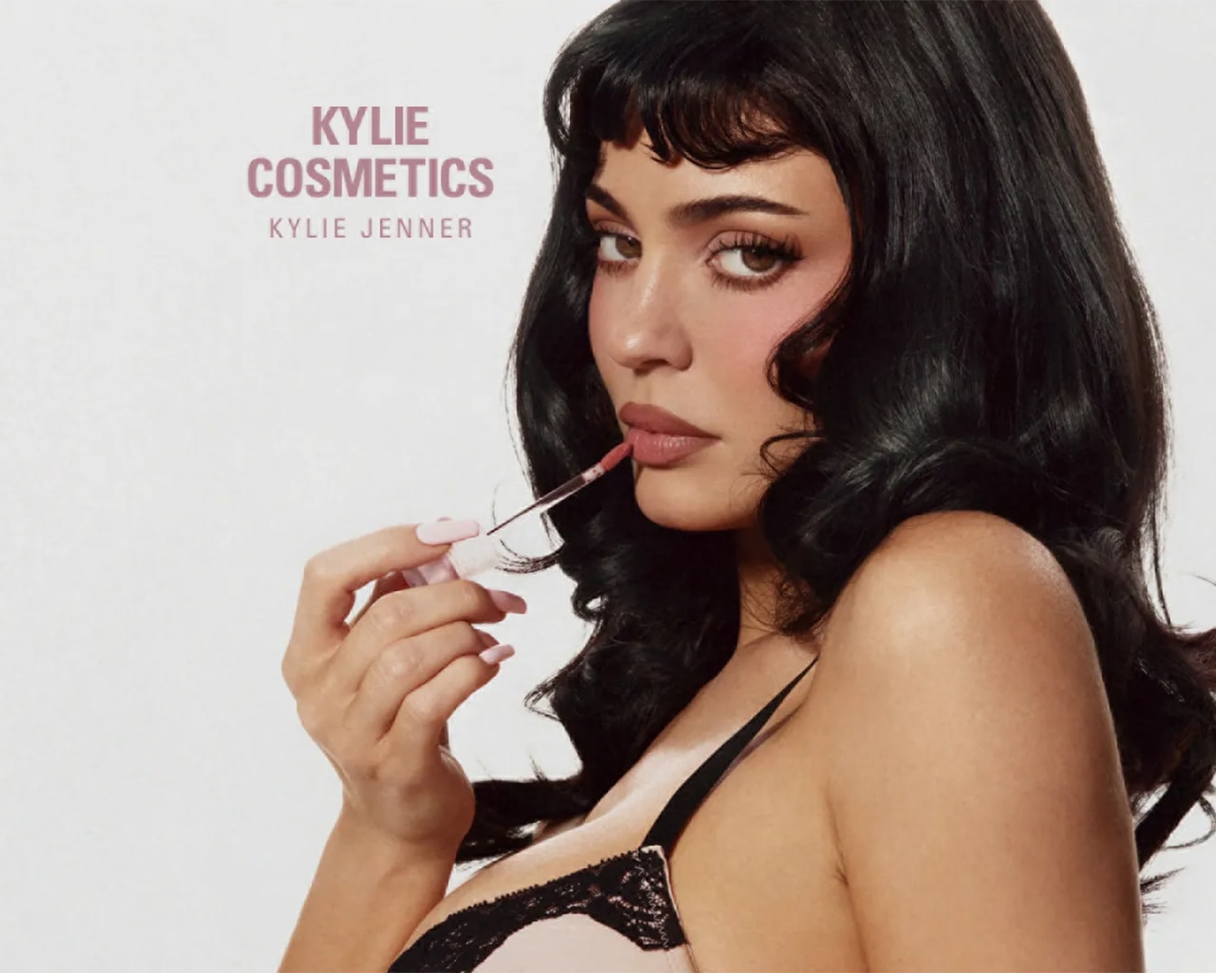 Kylie Jenner applica un lucidalabbra rosa chiaro, mettendo in risalto le sue labbra carnose e il trucco occhi, con la scritta "KYLIE COSMETICS KYLIE JENNER" in rosa chiaro su sfondo bianco.