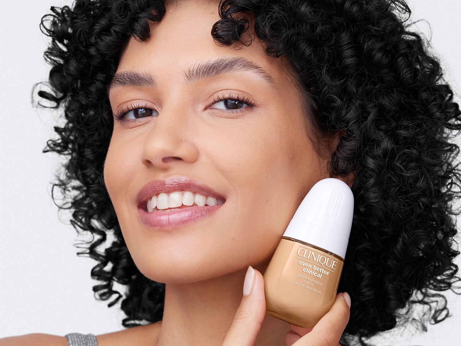 Uśmiechnięta kobieta o kręconych włosach trzyma w dłoni beżowy podkład Clinique Even Better Clinical Serum Foundation SPF 20 w butelce z białym aplikatorem.