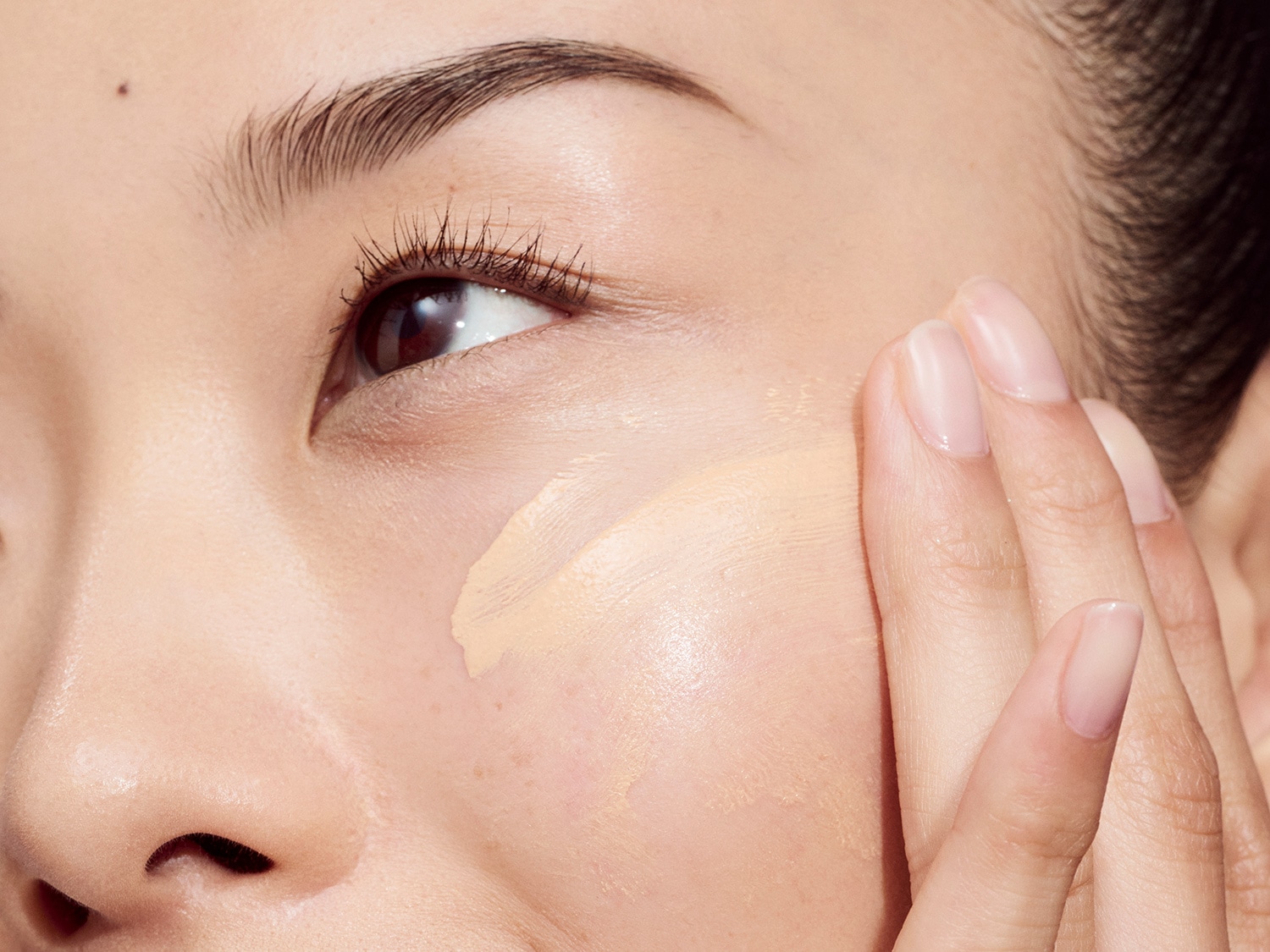 Na zbliżeniu widać kobietę nakładającą palcami beżowy podkład na policzek, aby uzyskać nieskazitelną cerę.