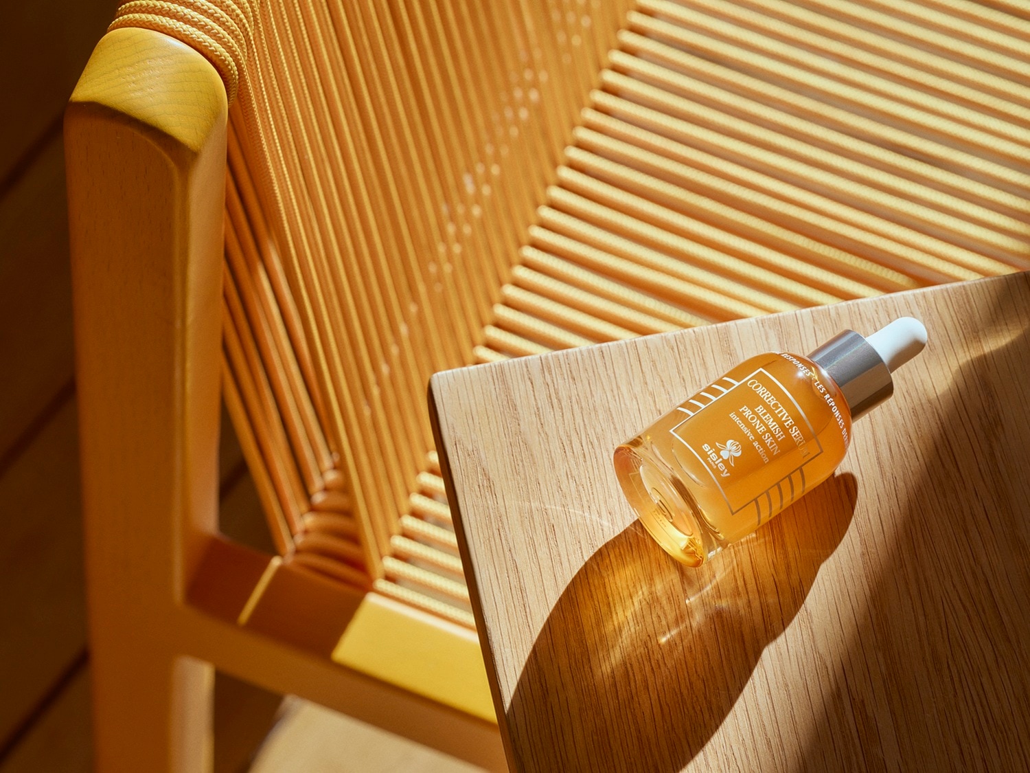 Buteleczka serum Sisley Corrective Serum Blemish Prone Skin o bursztynowym kolorze, z białą zakrętką i srebrnym aplikatorem, leży na drewnianym blacie obok żółtego krzesła.