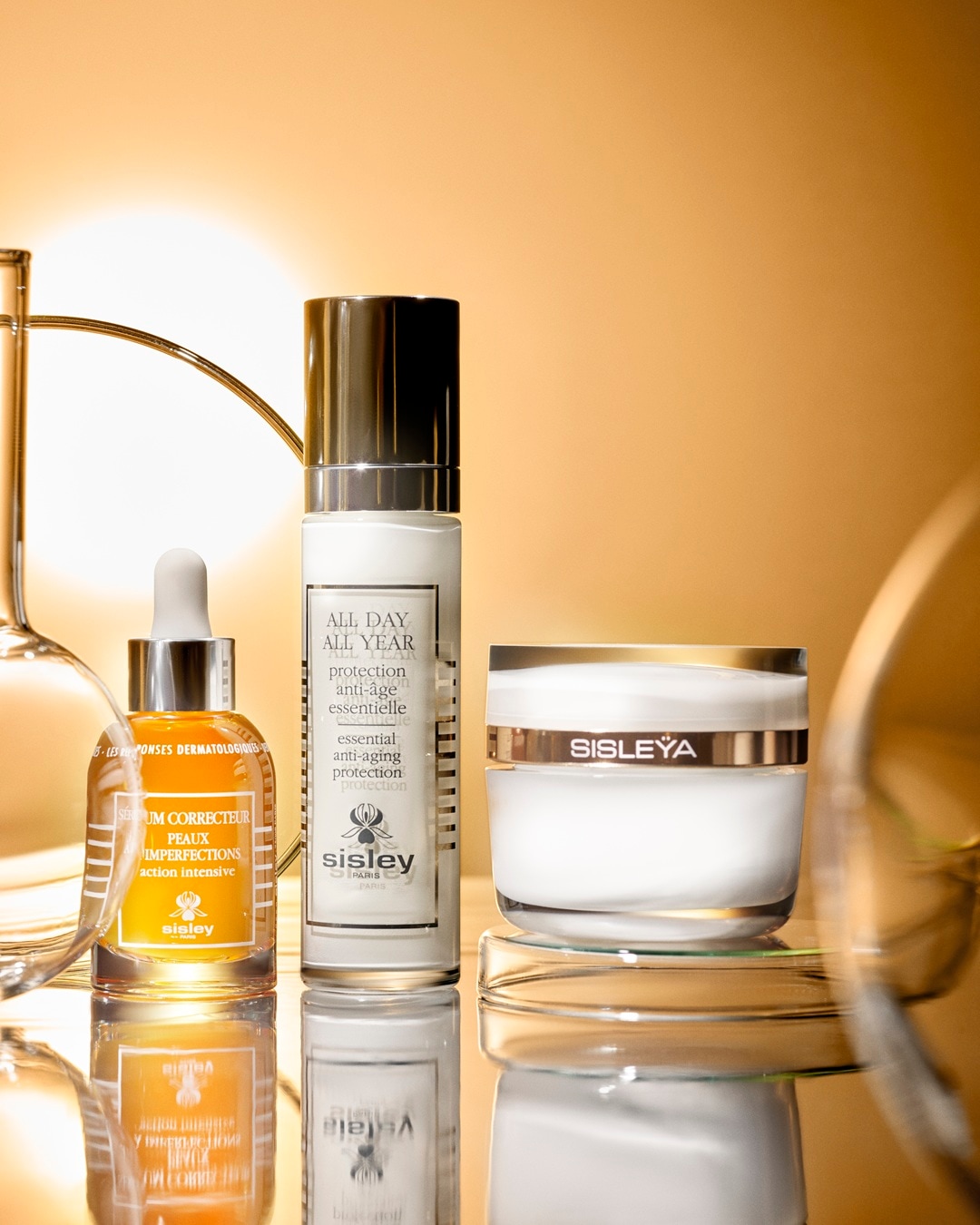 Trzy produkty Sisley Paris, w tym serum korygujące niedoskonałości w pomarańczowej butelce z zakraplaczem, biała butelka z pompką „ALL DAY ALL YEAR” oraz biały słoiczek kremu „SISLEYA”, stoją na błyszczącej powierzchni, odbijając się w niej.
