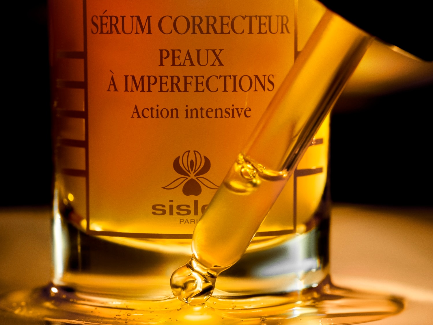Złociste serum do twarzy Sisley Paris SÉRUM CORRECTEUR PEAUX À IMPERFECTIONS z intensywnym działaniem, widoczne w przezroczystej butelce z pipetą, która nabiera płyn, a kilka kropel spływa po szklanej powierzchni.
